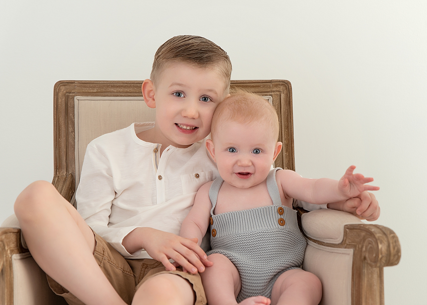 Two brothers sitting in a chair together. Both boys are blue eyes and are smiling. Photo by Rachel Brookes Photography.
